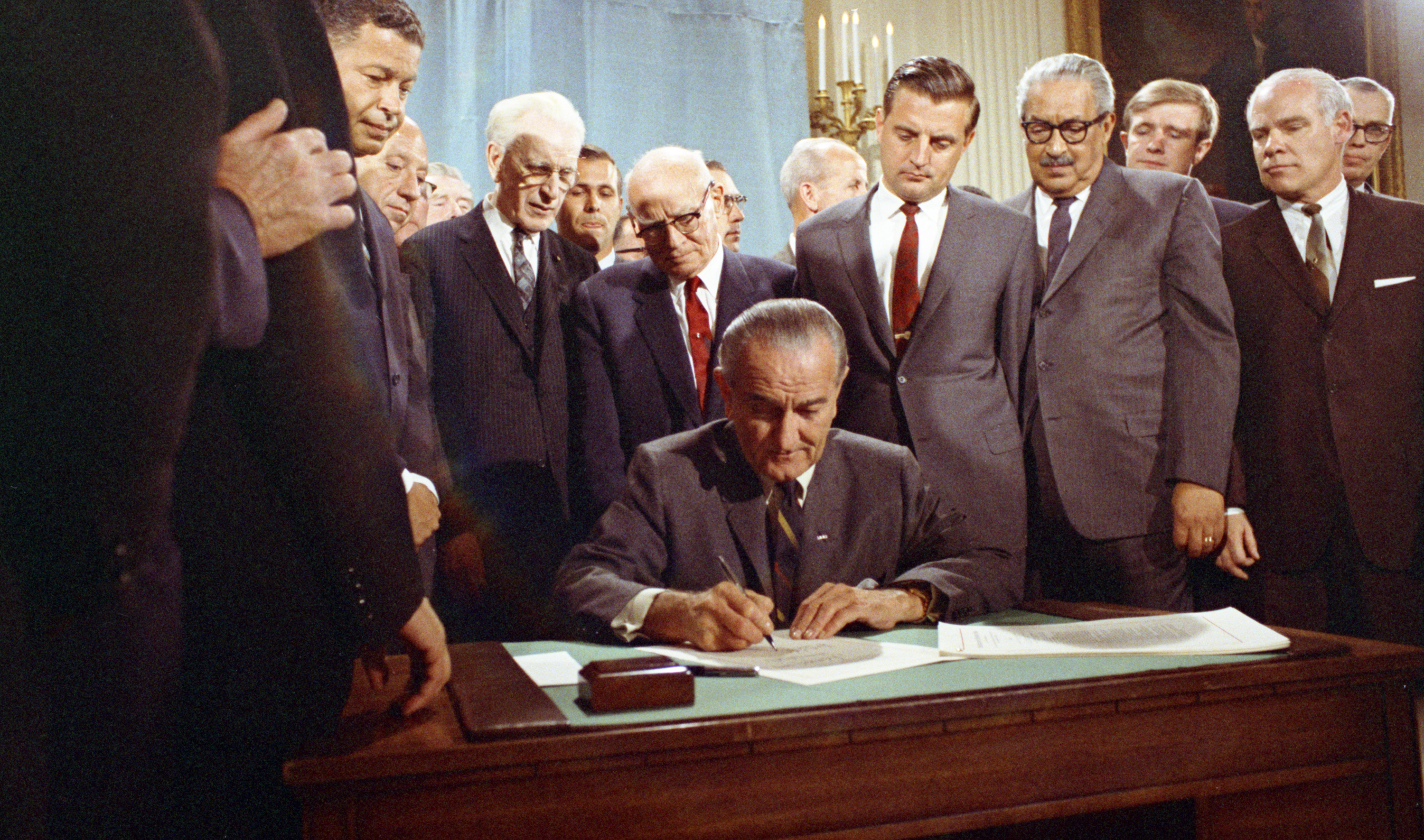 President Johnson, seated at desk, signs a piece of paper while a crowd of men in suits observe. 